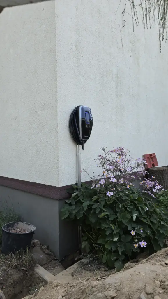 Schwarze Wallbox an einer Hauswand im Außenbereich, davor ein blühendes Beet